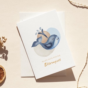 Op de afbeelding: Een wenskaart met een aquarel illustratie van een blauwe walvis met gesloten ogen, een waterspat en een lichtblauwe en bruine achtergrond. De kaart heeft de tekst "Alles Gute für die Elternzeit" in gouden letters.
