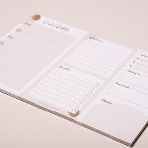 May include: A white weekly planner with a light brown watercolor design. The planner has sections for daily tasks, to-do list, weekly goals, intention, and focus.