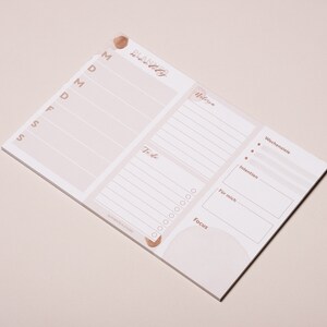 May include: A white weekly planner notepad with a pink and brown watercolor design. The notepad has sections for "To-do", "Focus", "For me", "Intentions", and "Wishes".