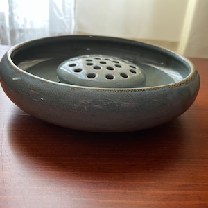 May include: A gray ceramic flower frog with a round, shallow bowl and a central, circular, perforated insert. The frog is designed to hold flower stems in place.