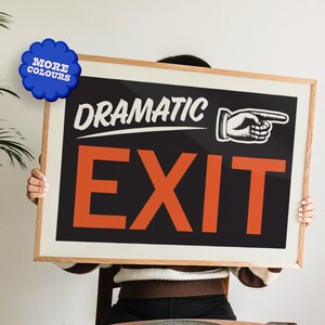 May include: A person is holding a wooden framed sign with the text "Dramatic Exit" in white lettering on a black background. The word "Exit" is in large orange letters with a black outline. A small black and white illustration of a hand pointing to the right is above the word "Exit".