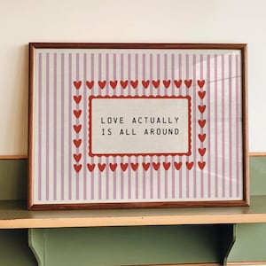 May include: A framed print with the text "LOVE ACTUALLY IS ALL AROUND" on a striped background with red hearts. The print is displayed on a vintage green and tan shelf, creating a charming, romantic aesthetic.
