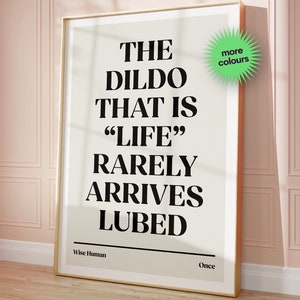 May include: A framed print with a white background and black text that reads "THE DILDO THAT IS "LIFE" RARELY ARRIVES LUBED". The text is in a bold, sans-serif font. The print is framed in a gold frame. The text "Wise Human" is in the bottom left corner and "Once" is in the bottom right corner. There is a green starburst sticker in the top right corner that says "more colours".