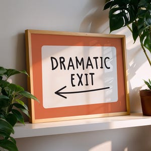 May include: A framed sign with the words "DRAMATIC EXIT" in black, with an arrow pointing left. The sign has a white background with an orange border and a wooden frame. The sign is on a white shelf with green plants.