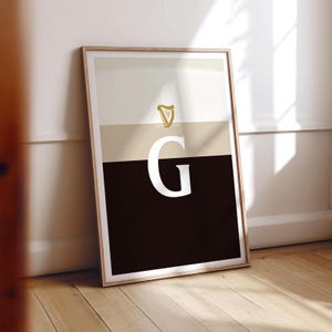 May include: A framed print with a black, white and gold colour scheme. The print features a stylised harp and the letter "G" in a bold, white font.