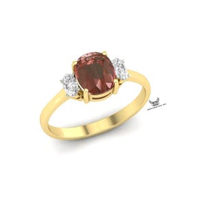 Puede incluir: Un anillo de oro con una gema grande, ovalada, de color marrón rojizo flanqueada por dos gemas claras más pequeñas. El anillo tiene un diseño clásico de tres piedras, sobre un fondo blanco.