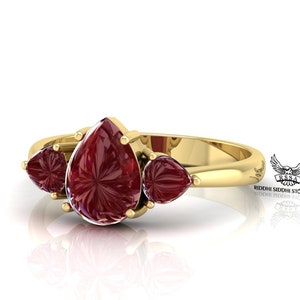May include: A gold ring featuring three pear-shaped, faceted, deep red gemstones. The central stone is larger than the two flanking stones. The ring has a polished finish and a simple band.