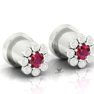 May include: A pair of silver-toned ear plugs featuring a flower-shaped front design. Each plug has a central round red gemstone surrounded by a halo of eight small, clear, round-cut stones set in a circular metal arrangement on a cylindrical base.