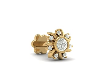 May include: A gold-colored nose stud in a sunburst design. The stud features a central, round, clear gemstone surrounded by smaller, clear gemstones. The design has a floral or star-like appearance, with curved petals or rays extending outwards.
