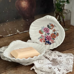 May include: Two white ceramic soap dishes shaped like seashells. The dish on the left contains a bar of soap. The dish on the right features a floral design in shades of pink, orange, blue, and green.