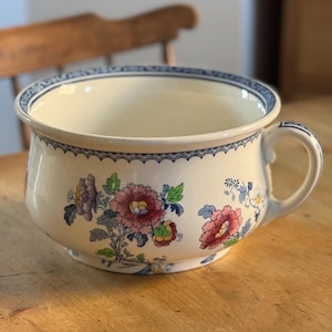 May include: A white ceramic chamber pot with a blue and white floral pattern. The pot has a handle on the side and a scalloped rim.