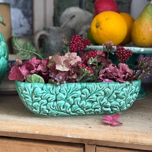 May include: A turquoise ceramic planter, filled with pink, red, and purple flowers. The planter has a detailed leaf pattern. Oranges and pears are visible in the background. The planter is on a wooden surface.