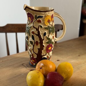 May include: A cream-coloured ceramic pitcher featuring a raised, embossed floral and tree branch design in shades of red, yellow, and green. The pitcher has a curved handle and a pouring spout, accompanied by two yellow lemons, an orange, and a red apple.