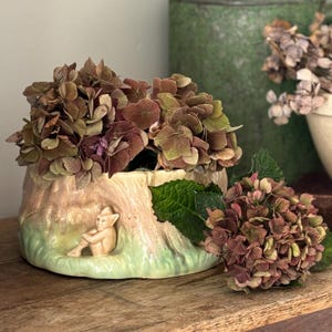 May include: A ceramic planter shaped like a tree stump with a small gnome figure sitting inside. The planter is decorated with a green glaze and has a brown and beige glaze to resemble bark. The planter is filled with dried hydrangeas.