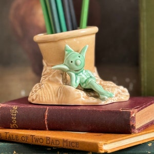 May include: A quirky ceramic pencil holder in the shape of a boot, featuring a light green elf figurine. The boot is tan, and the elf is light green. Green pencils are inside the boot. The holder sits on a stack of vintage books.