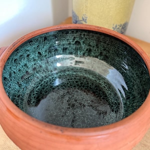 May include: A handcrafted terracotta bowl featuring a speckled teal interior and a reddish-brown rim. The bowl has a glossy finish and a rustic, artisanal appearance. The bowl is likely used for serving food or as a decorative item.