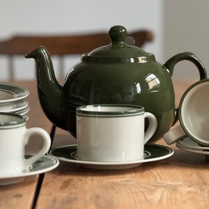 May include: A green ceramic teapot with a white and green striped teacup and saucer. The teapot has a round body and a spout on the side. The teacup and saucer are sitting on a wooden table.