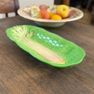 May include: An elongated, oval-shaped ceramic serving dish in a vibrant green hue, featuring a raised, moulded relief of a celery stalk extending along its centre. The dish has a glossy finish and textured edges that mimic the appearance of leaves.