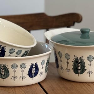 May include: Set of three white ceramic bowls with a floral design in shades of blue, green, and black. The bowls have a scalloped edge and are stacked on top of each other. The largest bowl has a lid.
