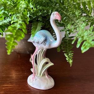 May include: A ceramic flamingo figurine with a pink beak and legs, a light blue body, and a white neck. The flamingo is perched on a decorative base with green and pink accents. The figurine is a decorative item.