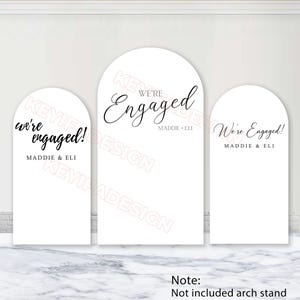 May include: Three white arched signs featuring black text. The left sign reads "we're engaged! MADDIE & ELI". The centre sign reads "WE'RE Engaged MADDIE + ELI". The right sign reads "We're Engaged! MADDIE & ELI". The signs are flat, smooth, and rectangular with rounded tops.