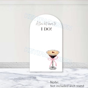 以下が含まれることがあります： 「A tini bit closer to I DO!」のテキストが入った白いアーチ型のサイン。ピンクのリボンが付いたマティーニグラスの水彩イラストが下にあります。サインは大理石の表面にあります。
