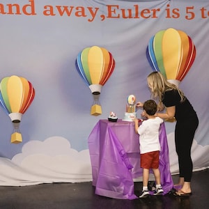 May include: A child blows out candles on a birthday cake decorated with hot air balloons. The cake is on a purple tablecloth in front of a backdrop with a blue sky, white clouds, and hot air balloons. The backdrop has the text "and away, Euler is 5 to"