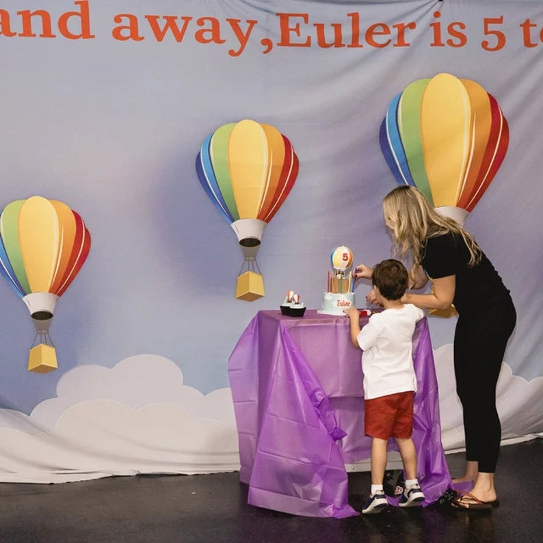 May include: A child blows out candles on a birthday cake decorated with hot air balloons. The cake is on a purple tablecloth in front of a backdrop with a blue sky, white clouds, and hot air balloons. The backdrop has the text "and away, Euler is 5 to"