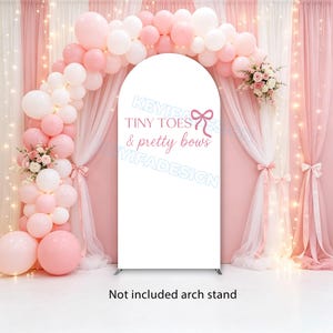 May include: A white arched backdrop cover featuring the pink text Tiny Toes & pretty bows with a bow graphic. The cover is displayed with a balloon arch in shades of pink and white, accented by floral arrangements and sheer fabric curtains.