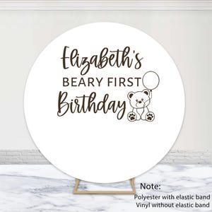 May include: White round backdrop with the text "Elizabeth's Beary First Birthday" and a cartoon bear holding a balloon.
