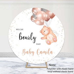 Op de afbeelding: Een ronde witte achtergrond met gouden glitter en een teddybeer die roze en gouden ballonnen vasthoudt. De tekst "WE CAN bearly WAIT Baby Camila" staat op de achtergrond.