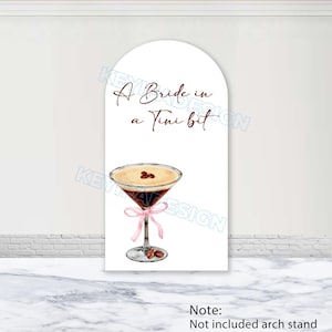 Könnte beinhalten: Weißes, bogenförmiges Schild mit dem Text "A Bride in a Tini bit" über einer Aquarellillustration eines Martinis. Der Martini hat ein rosa Band und Kaffeebohnen. Auf dem unteren Teil des Schildes steht "Note: Not included arch stand."
