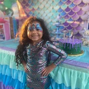 May include: A young girl wearing a mermaid costume with iridescent scales and a mermaid-themed face paint. She is standing in front of a mermaid-themed cake with purple frosting and green fondant.