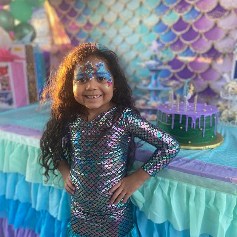 May include: A young girl wearing a mermaid costume with iridescent scales and a mermaid-themed face paint. She is standing in front of a mermaid-themed cake with purple frosting and green fondant.