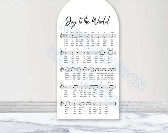 May include: An arched white sign featuring the black printed text Joy to the World at the top, followed by musical notation and lyrics for the Christmas carol. The design includes staff lines, notes, and chord symbols on a clean white background.
