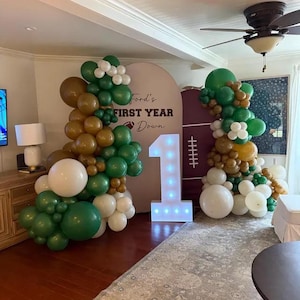 May include: A first birthday party decoration set featuring a large illuminated number one, two balloon arches in shades of green, gold, and white, and a backdrop with the text Ford's FIRST YEAR Down and a football graphic.