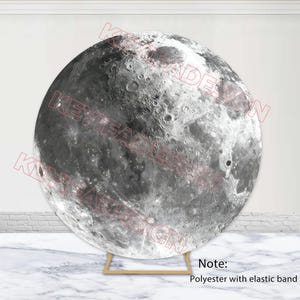 May include: A large, round, grayscale backdrop featuring a detailed depiction of the moon's surface, complete with craters and textures. The backdrop is supported by a simple, gold-colored stand. The text "KEYIFADE DESIGN" is visible on the backdrop.