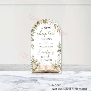 May include: An arched sign featuring the text A NEW chapter BEGINS WELCOME TO Emily's BRIDAL SHOWER. The design includes watercolor illustrations of green foliage, white flowers, and an open book at the bottom. The sign has a white background.