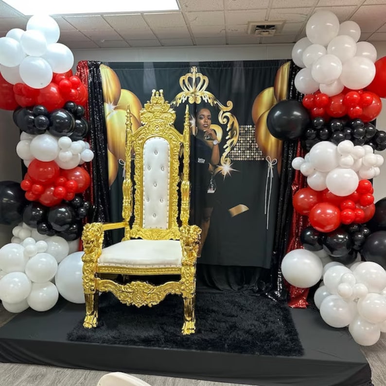 May include: A gold throne chair with a white cushion sits on a black platform in front of a black backdrop with gold accents. The backdrop features a crown and the words "Queen" and "Boss".  The chair is decorated with gold trim and has a white cushion.  There are red and black balloons on either side of the chair.