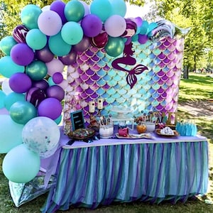 May include: A mermaid-themed party table with a backdrop featuring a mermaid tail and purple and blue scales. The table is decorated with a purple and blue tulle tablecloth, balloons, and various treats.