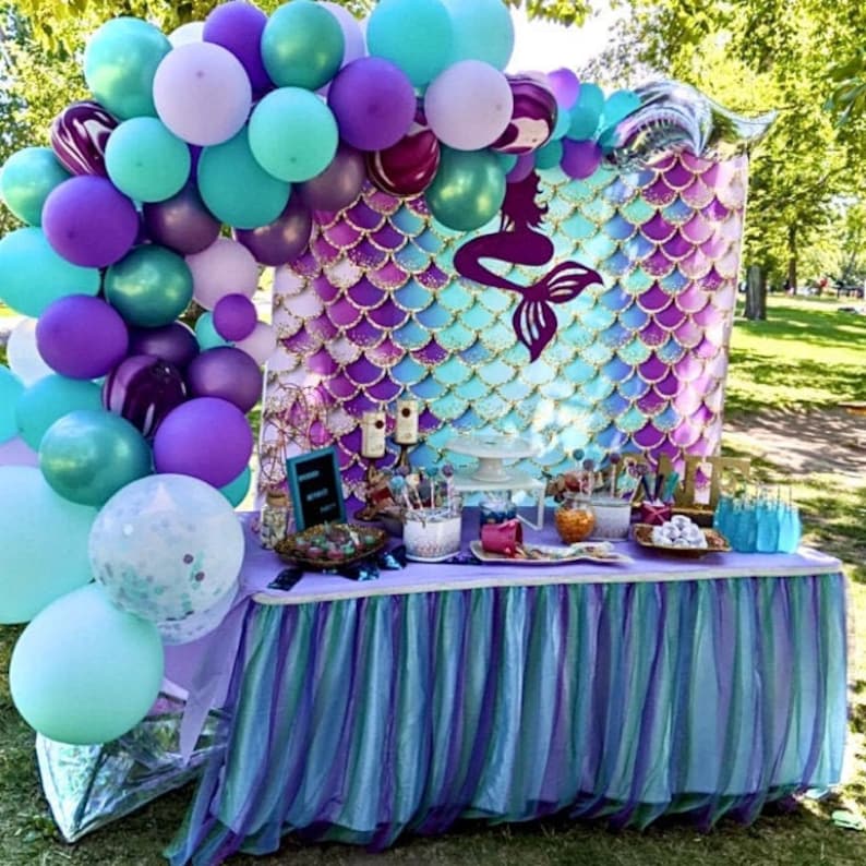 May include: A mermaid-themed party table with a backdrop featuring a mermaid tail and purple and blue scales. The table is decorated with a purple and blue tulle tablecloth, balloons, and various treats.