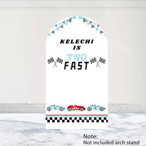 May include: A white arch-shaped sign with the text "Kelechi is Two Fast" in black and blue. The sign features racing flags, a checkered border, and illustrations of race cars in blue and red. A banner of flags decorates the top.