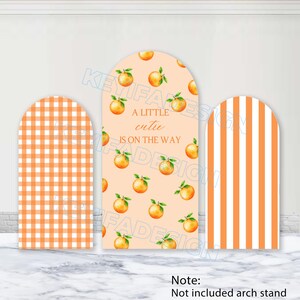 Puede incluir: Tres letreros en forma de arco con patrones naranjas y blancos. El letrero central tiene el texto "A LITTLE cutie IS ON THE WAY" con ilustraciones de frutas naranjas. Los otros letreros tienen un patrón de cuadros y rayas.