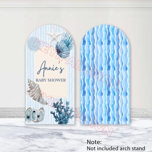 Peut inclure: Deux panneaux de baby shower en forme d'arche. L'un présente un fond à rayures bleues et blanches avec des coquillages et le texte "Annie's BABY SHOWER". L'autre panneau a un motif ondulé bleu. Les panneaux sont décorés d'illustrations de coquillages et de coraux.