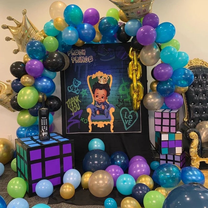 May include: A baby shower decoration with a black backdrop featuring the words "Fresh Prince" and a cartoon image of a baby in a crown sitting on a throne. The backdrop is decorated with graffiti-style artwork. The decoration also includes a balloon arch with purple, green, blue, and gold balloons. There are also two Rubik's Cube-style boxes and a gold throne.