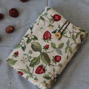 Könnte beinhalten: Eine rechteckige Hülle mit Erdbeermuster. Die Hülle hat einen beigefarbenen Hintergrund mit roten Erdbeeren, grünen Blättern und weißen Blüten. Ein Holzknopf und ein schwarzes Gummiband sichern die Hülle. Kastanien sind auf einer blauen Oberfläche verstreut.