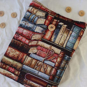 Puede incluir: Una funda de tela rectangular con un patrón tejido de libros apilados en tonos rojo, azul, amarillo y marrón. La funda se asegura con un cordón elástico negro que se engancha sobre un botón de madera de color claro en la parte frontal.
