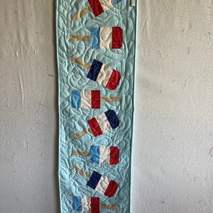 May include: A blue quilted table runner with a row of red, white, and blue popsicle-shaped fabric appliques. The runner has a scalloped edge and a white label that says "SMALL".