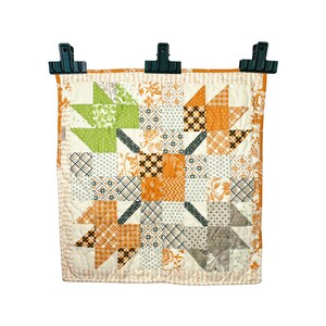 May include: A small, orange, green, and white patchwork quilt with a starburst design. The quilt is hanging from three black clips against a white background.