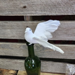 May include: A white dove figurine perched on top of a green glass bottle. The bottle is sitting on a wooden surface.